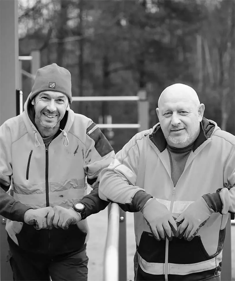 Two men in work attire smiling during playground maintenance.